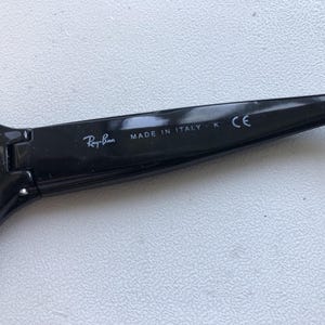 May include: Close-up of a black Ray-Ban sunglasses arm. The text "Ray-Ban" is visible, along with "MADE IN ITALY" and "CE". The sunglasses arm is glossy black.