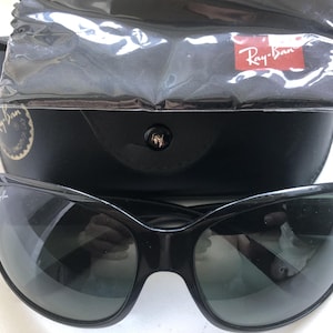 May include: Black sunglasses with a curved frame, resting on top of a black case. A cleaning cloth and a red Ray-Ban logo are visible. The sunglasses have dark lenses.