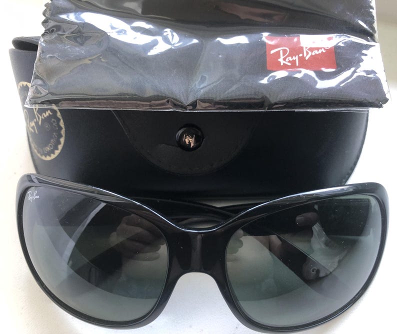May include: Black sunglasses with a curved frame, resting on top of a black case. A cleaning cloth and a red Ray-Ban logo are visible. The sunglasses have dark lenses.