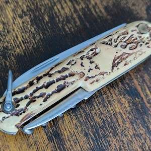 May include: A folding pocket knife with a textured, cream-colored handle featuring dark brown, irregular grooves. The knife includes a silver-toned metal blade, a small metal bail loop at one end, and a corkscrew tool folded into the handle side.