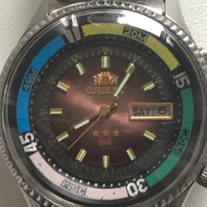 May include: A stainless steel Orient watch with a brown face, luminous hands, and a day-date display. The bezel features colored markings in blue, yellow, green, and pink. The watch has a metal band and is designed for diving.
