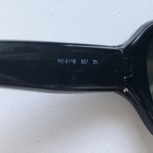 May include: Close-up of black sunglasses with dark lenses. The arm of the glasses has the numbers "RB 4118 601 3N" printed on it. The sunglasses have a rectangular shape.