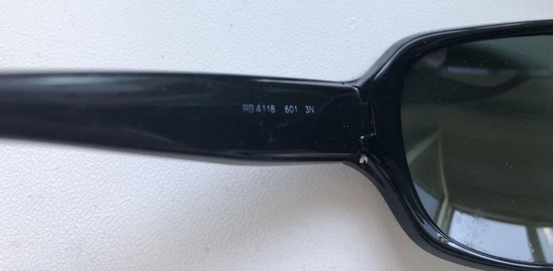 May include: Close-up of black sunglasses with dark lenses. The arm of the glasses has the numbers "RB 4118 601 3N" printed on it. The sunglasses have a rectangular shape.