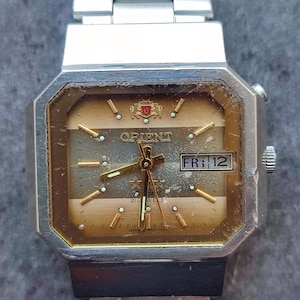 May include: Vintage Orient watch with a silver-tone stainless steel band and case. The watch face is a gold color with a day and date display. The watch has a rectangular shape with a textured dial and luminous hands.