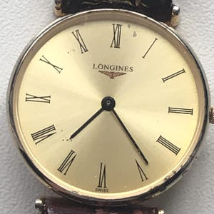 May include: A gold Longines wristwatch with a round face, black Roman numerals and hands. The watch has a gold bezel and a brown leather strap. The word "SWISS" is printed at the bottom of the watch face.