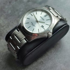 Op de afbeelding: Een zilveren Orient-horloge met een witte wijzerplaat en zilveren wijzers. Het horloge heeft een datumvenster op 3 uur en de woorden "Orient" en "Water Resist 100m" staan op de wijzerplaat.