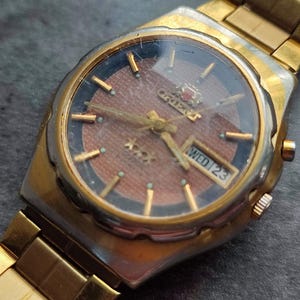 May include: A gold-toned wristwatch with a brown face and black accents. The watch face features a day and date display, as well as the brand name "Orient".