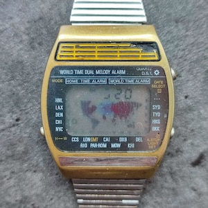 May include: Vintage gold-toned digital watch with a silver band. The watch face displays the time and world time zone abbreviations. Features include alarm and date functions. The text "WORLD TIME DUAL MELODY ALARM" is visible.