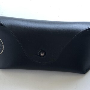 May include: Black leather sunglasses case with a snap closure. The case has a rounded shape and features the Ray-Ban logo in a silver circle on the front. The case is designed to protect eyewear.