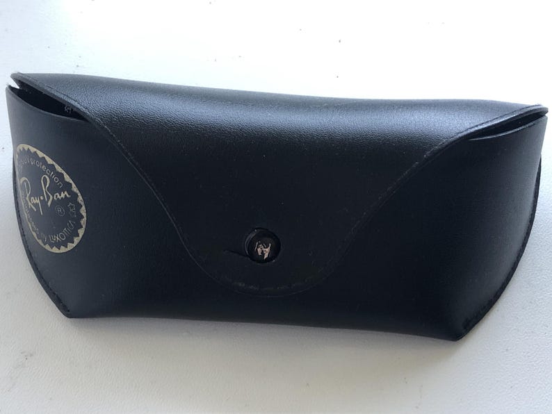 May include: Black leather sunglasses case with a snap closure. The case has a rounded shape and features the Ray-Ban logo in a silver circle on the front. The case is designed to protect eyewear.