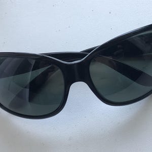 May include: Black sunglasses with a curved frame and dark lenses. The sunglasses have a glossy finish and a small logo on the left side. The lenses are reflective, and the overall design is sleek and modern.
