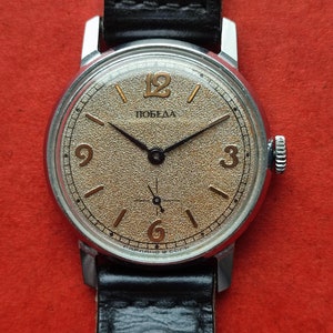 Könnte beinhalten: Eine Vintage-Armbanduhr mit silberfarbenem Gehäuse und einem strukturierten, beigefarbenen Zifferblatt. Das Zifferblatt hat goldfarbene Ziffern und Zeiger, mit dem Wort "Победа". Das Armband ist aus schwarzem Leder.