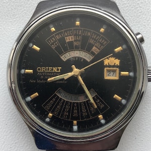 May include: A black Orient automatic wristwatch with a gold-tone face and hands. The watch features a multi-calendar display with day, date, and month indicators. The watch face also includes the text "Orient Automatic 21 Jewels New Multi-Calendar".
