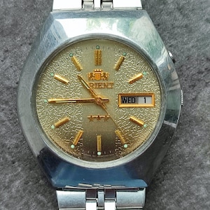 May include: A silver wristwatch with a gold face and a gold-colored logo that says "Orient" on it. The watch has a day and date window that says "WED 1".