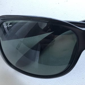 May include: Close-up of black sunglasses with the Ray-Ban logo in white on the upper left corner. The lenses are dark, reflecting the surrounding environment. The frame is thick and curved.