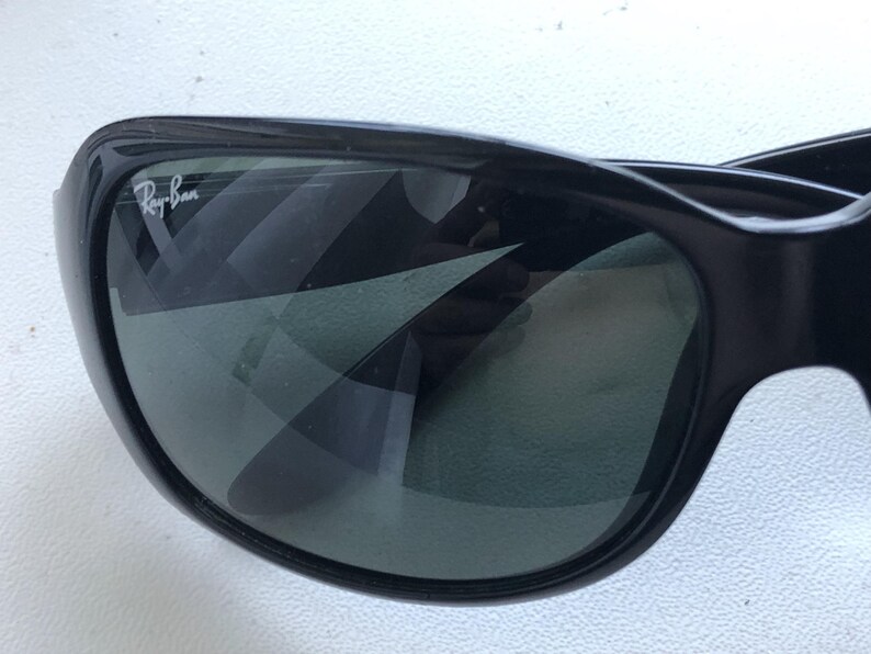 May include: Close-up of black sunglasses with the Ray-Ban logo in white on the upper left corner. The lenses are dark, reflecting the surrounding environment. The frame is thick and curved.