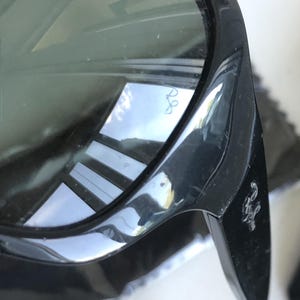May include: Close-up of black sunglasses with dark lenses. The frame is glossy and reflects the surrounding environment. The Ray-Ban logo is visible on the lens and the temple.