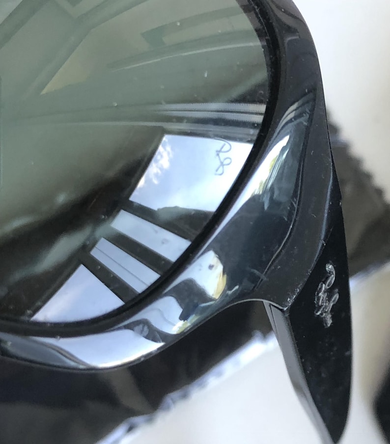 May include: Close-up of black sunglasses with dark lenses. The frame is glossy and reflects the surrounding environment. The Ray-Ban logo is visible on the lens and the temple.