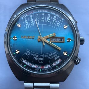 May include: Vintage Orient 21 Jewels watch with a blue dial and a stainless steel band. The watch face displays the day, date, and month, with gold-tone hands and markers. The watch has a calendar function and is marked "Japan".