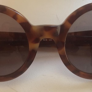 Puede incluir: Gafas de sol redondas con montura de carey en tonos marrón y negro y lentes oscuras. La montura presenta pequeños detalles metálicos en los bordes exteriores y un puente que conecta los dos aros circulares de la estructura.