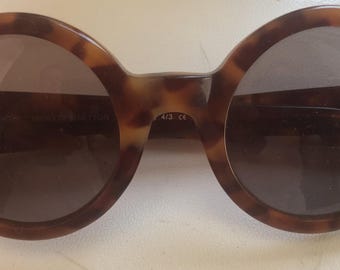 Puede incluir: Gafas de sol redondas con montura de carey en tonos marrón y negro y lentes oscuras. La montura presenta pequeños detalles metálicos en los bordes exteriores y un puente que conecta los dos aros circulares de la estructura.