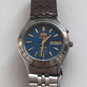 May include: A silver wristwatch with a blue face and gold accents. The watch face has the words "Orient Crystal 21 Jewels" and the day of the week "TUE" displayed. The watch has a silver metal band.