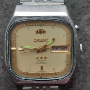 May include: A silver Orient watch with a white face and gold accents. The watch has a day and date display and says "Crystal 21 Jewels".