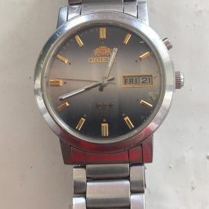 May include: A silver Orient watch with a black face and gold accents. The watch has a date window that displays "FRI 21".