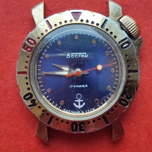 Puede incluir: Un reloj de pulsera dorado con una esfera azul y una aguja de segundos roja. La esfera del reloj tiene la palabra "Восток" y el texto "17 КАМНЕЙ" impreso en ella. El reloj tiene un bisel dorado con números negros y un triángulo rojo en la parte superior.