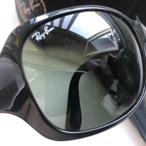 May include: Black Ray-Ban sunglasses with a glossy finish. The lenses are a gradient gray, and the Ray-Ban logo is visible on the upper left lens. The sunglasses reflect the surrounding environment.