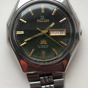 May include: A vintage Ricoh automatic wristwatch with a silver case and strap. The watch face is dark green with gold hands and markers. The day and date are displayed at the 3 o'clock position. The watch reads "21 Jewels Automatic".