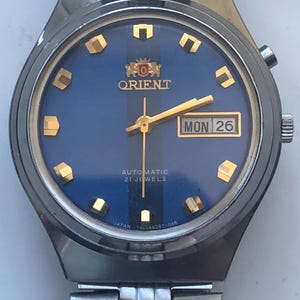 May include: A vintage Orient automatic wristwatch with a blue dial and gold-tone hands and markers. The watch face displays "Orient," "Automatic 21 Jewels," and "Mon 26." The stainless steel band and case complete the classic design.