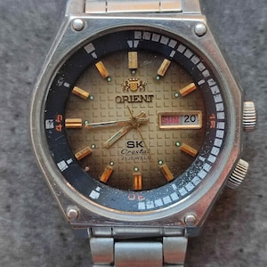 May include: A silver Orient SK Crystal watch with a black face and gold accents. The watch has a date window that displays "SUN 20" and a 24-hour subdial. The watch is on a silver metal band.