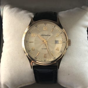 Puede incluir: Reloj de pulsera automático Adriatica dorado con esfera color crema, correa de cuero negro y visualización de la fecha. El reloj presenta agujas e indicadores horarios dorados. El reloj se muestra sobre una almohada blanca.