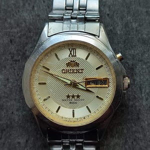 May include: A silver Orient watch with a white face, gold hands, and a date window. The watch has a silver metal band and says "Water Resist 50m" on the face.