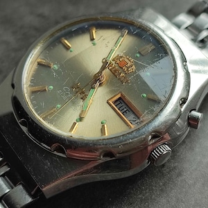 May include: A silver wristwatch with a silver metal band. The watch face is silver with black markings and a day and date window. The watch has a green second hand and a gold crown.