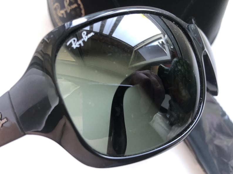 May include: Black Ray-Ban sunglasses with a glossy finish. The lenses are a gradient gray, and the Ray-Ban logo is visible on the upper left lens. The sunglasses reflect the surrounding environment.