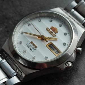 May include: A silver wristwatch with a white face and gold accents. The watch has a date window and a day of the week window. The watch face says "Crystal 21 Jewels" and "King Orient".