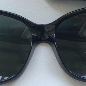 May include: Black sunglasses with dark green lenses. The frames have a rounded, slightly cat-eye shape. The lenses reflect the surrounding environment. The sunglasses are a classic accessory.