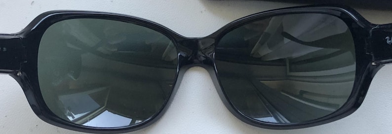 May include: Black sunglasses with dark green lenses. The frames have a rounded, slightly cat-eye shape. The lenses reflect the surrounding environment. The sunglasses are a classic accessory.