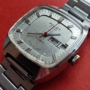Puede incluir: Un reloj de pulsera plateado con una esfera cuadrada. La esfera del reloj tiene marcas negras y un fondo blanco. El reloj tiene una correa de metal plateado y dice "Poljot 23 Jewels Automatic Made in USSR" en la esfera. El día de la semana se muestra como "WED" y la fecha es "27".