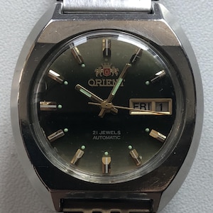 May include: An Orient automatic wristwatch with a dark green dial, silver-tone hands, and hour markers. The watch features a day-date window at the three o'clock position displaying FRI 1, a stainless steel case, and a matching multi-link metal bracelet.