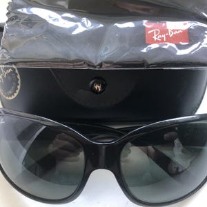 May include: Black Ray-Ban sunglasses with a curved frame and dark lenses. The sunglasses are displayed with a black case, a cleaning cloth, and a Ray-Ban logo tag.