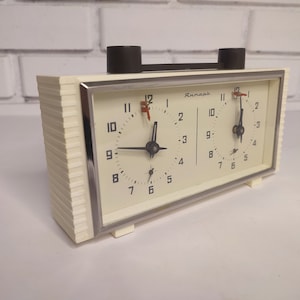 May include: A white analogue chess clock with two faces, each with black numbers and hands. The clock is labelled "Aurant" and has a silver trim.