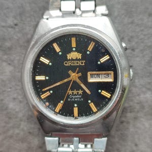 May include: A silver Orient watch with a black face and gold accents. The watch has a day and date window, and the face says "Crystal 21 Jewels".