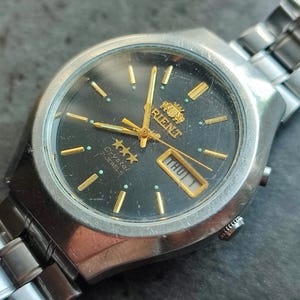 May include: A silver Orient Crystal 21 Jewels watch with a black face, gold hands, and a day of the week display. The watch has a silver metal band.