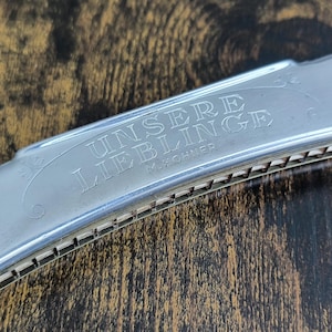 May include: A silver-colored metal harmonica with the engraved text "UNSERE LIEBLINGE" and "M. HOHNER" on the top cover plate. The musical instrument features a series of rectangular air holes along the side and the letter "C" on the right end.