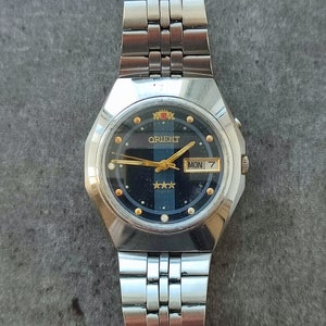 May include: A silver Orient wristwatch with a black face and gold accents. The watch has a day and date window and a silver metal band.