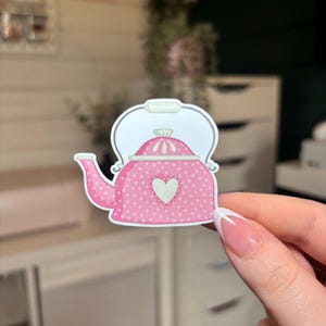 May include: A pink teapot sticker with white polka dots and a white heart design. The sticker has a white outline and a handle. The teapot is a cute, whimsical design.