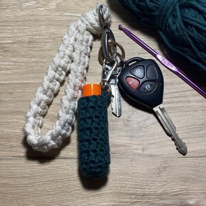 May include: A keychain featuring a cream-colored macrame wristlet attached to a metal clasp, a dark green crocheted lip balm holder with an orange cap, a metal key, and a black car key fob, alongside a ball of dark green yarn and a purple crochet hook.